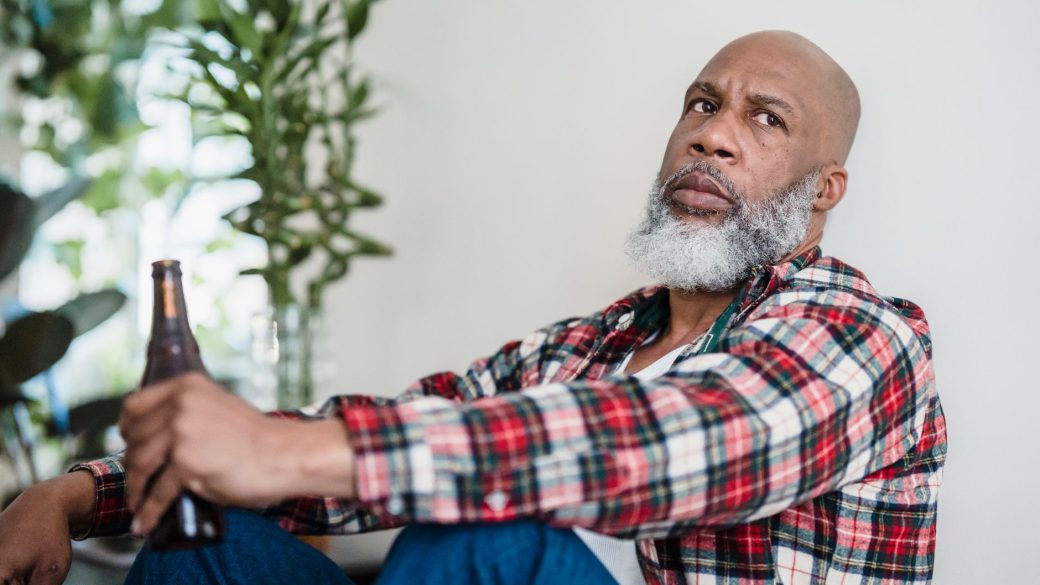 Bearded man in plaid shirt, sitting and holding a beer indoors.