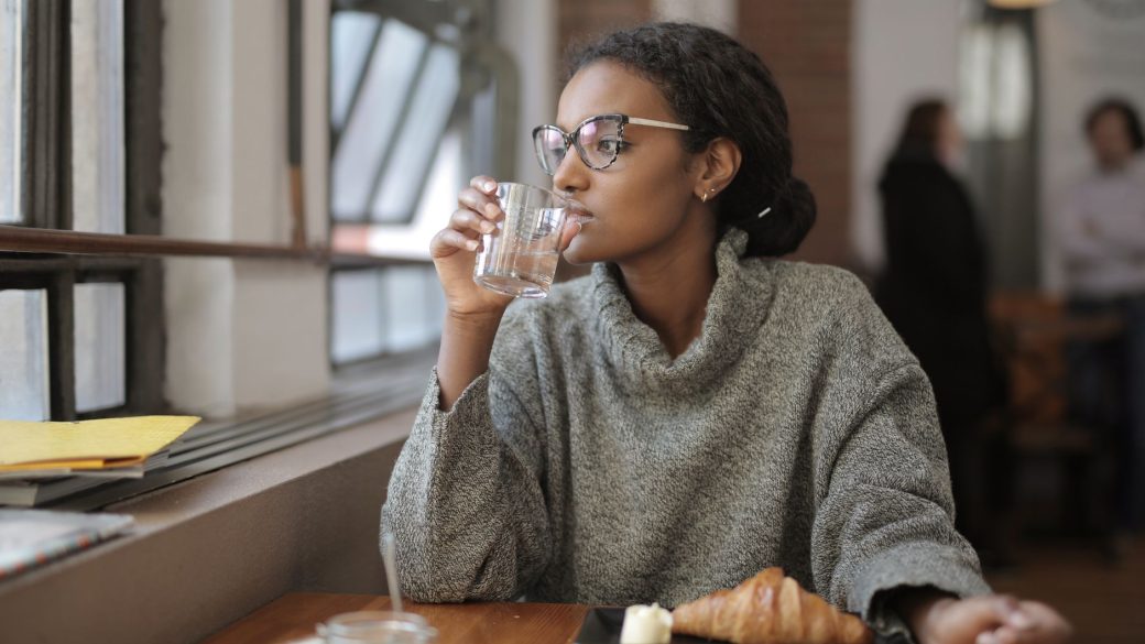 African American woman sipping water with a croissant at a cafe. Cozy indoor setting.