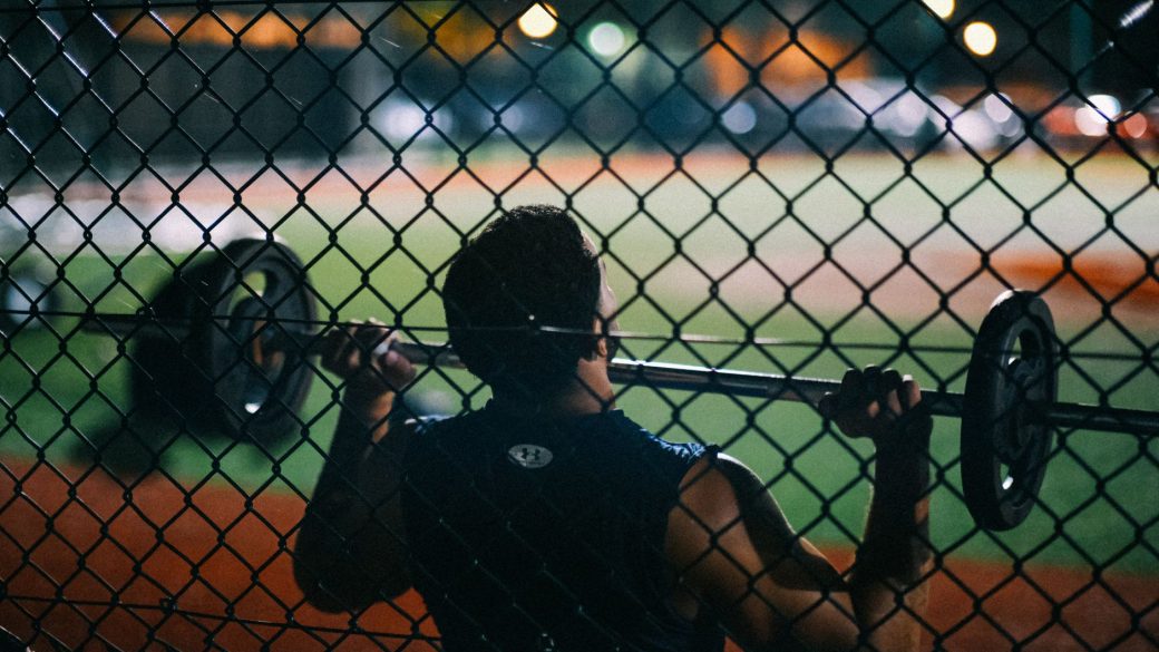 A man lifting weights behind a chain fence at night, showcasing strength and fitness.
