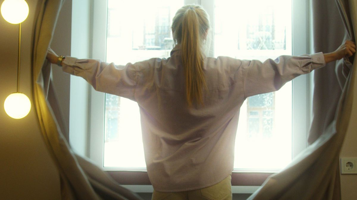 Woman opening bedroom curtains for morning light to reset her sleep rhythm