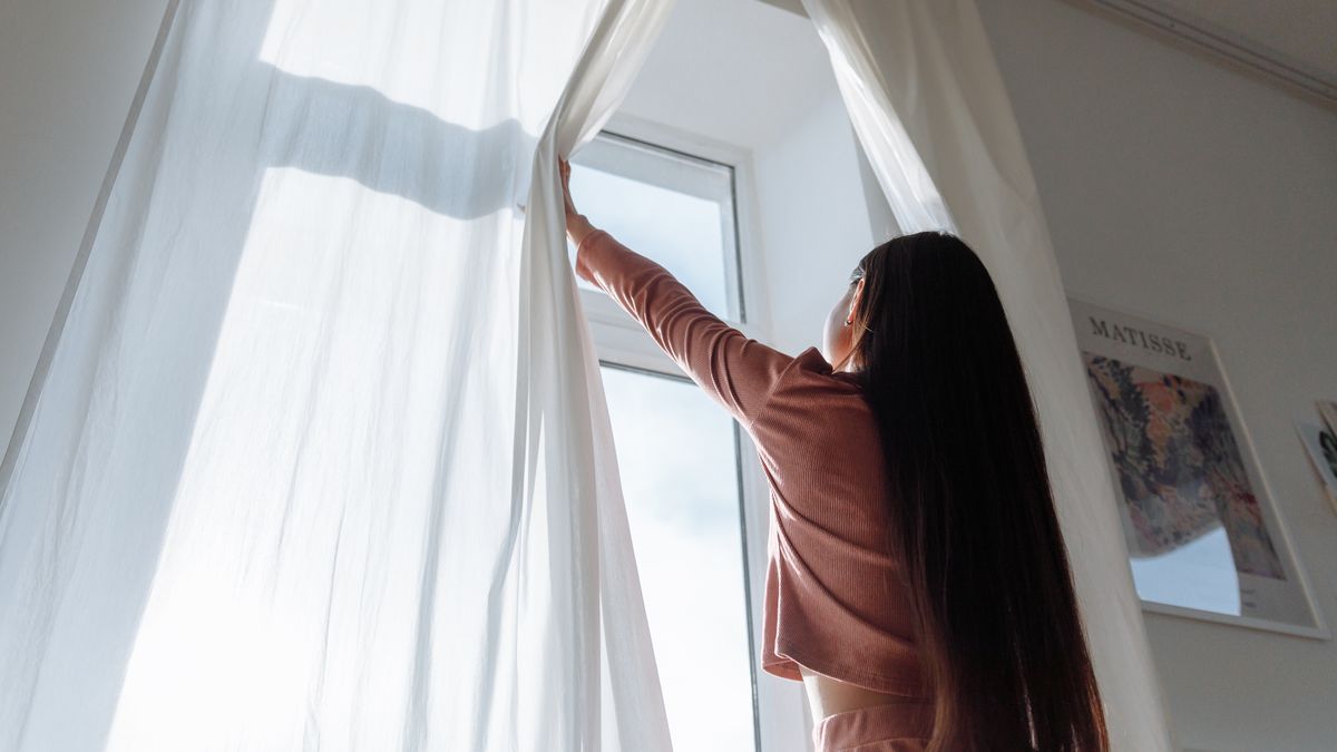Woman opening curtains to morning sunlight, which anchors the body clock and evening melatonin